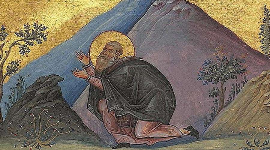 Hilarion, portrayed as a bearded man with a halo, kneels in prayer, facing left. A blue-and-purple mountain fills the background, and the sides of the image depict stylized desert plants.