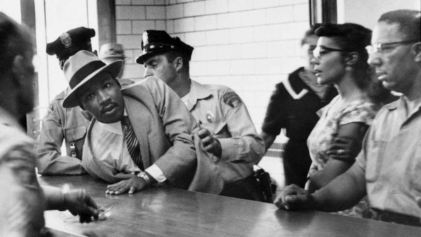 Dr. King, wearing a tan suit and hat, is being pushed across a counter by two white police officers. He is looking at a white official behind the counter. Mrs. King, wearing glasses and a light patterned dress, is standing with at least two other Black observers: one man and another who is invisible except for a ringed left hand on Mrs. King’s upper arm. Two indistinct figures are walking in the background.